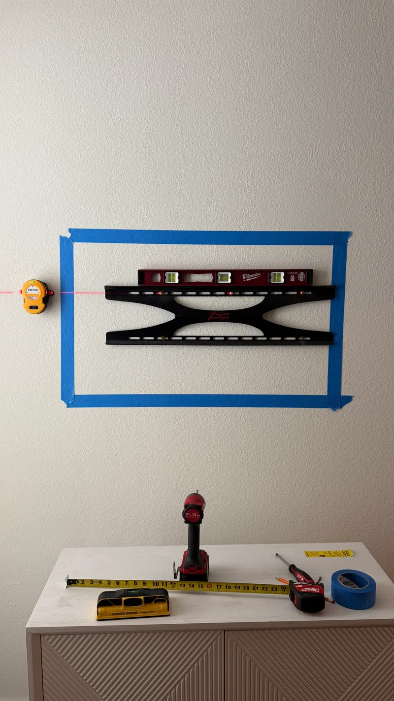 A TV bracket is positioned within a taped layout, with a spirit level and laser reference.