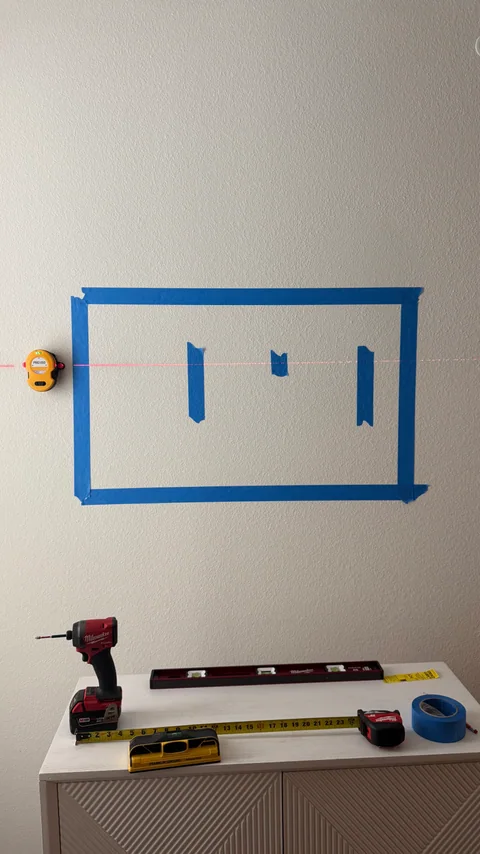Painter’s tape marks a proposed screen position above the cabinet, with a laser reference and tools below.